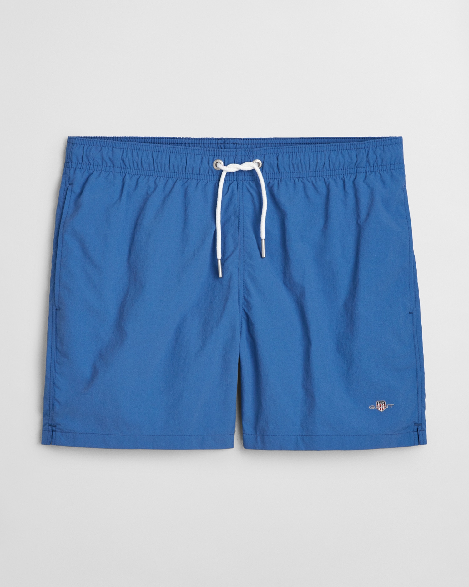 Blue swim shorts with an elastic waistband and a white drawstring. A small embroidered logo is visible on one leg. The fabric appears lightweight and suitable for swimming. Shorts are laid flat on a plain background.