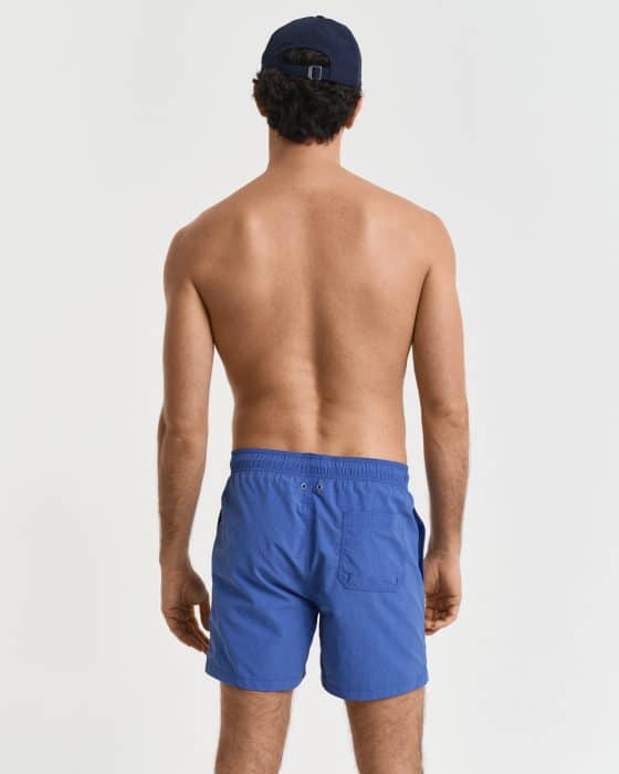 A man wearing a navy cap and blue shorts is standing with his back to the camera. He is shirtless and in a plain studio setting with a white background.