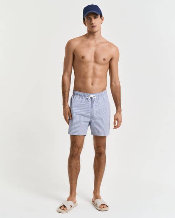 A man stands against a neutral background wearing a navy cap, light blue swim shorts with a drawstring, and white slide sandals. He is shirtless, with a relaxed posture and neutral expression.