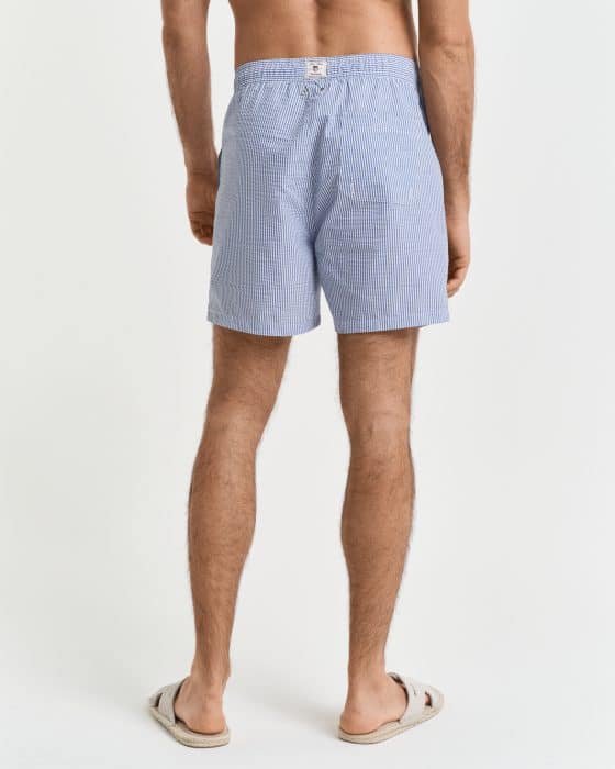 Rear view of a person wearing light blue checkered shorts and beige slippers. They are standing against a plain white background, with visible partial back and legs. The shorts are loose-fitting with an elastic waistband.