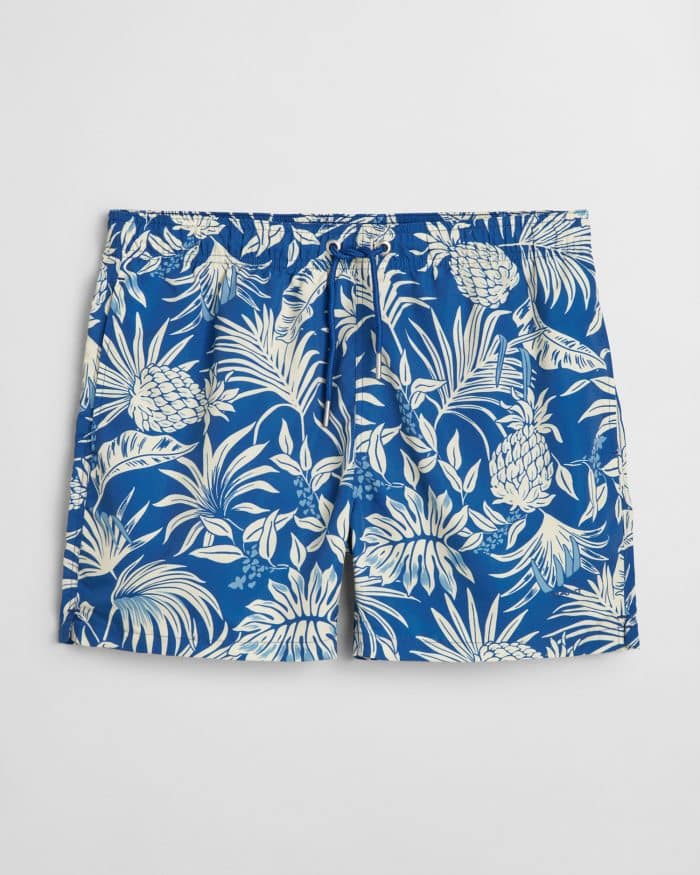 Blue swim shorts featuring a tropical pineapple and leaf pattern in white. The shorts have an elastic waistband with a drawstring.