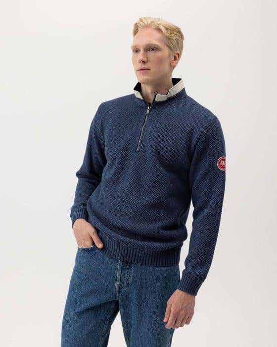 A person with short blonde hair wearing a blue zip-up sweater with a white collar and a red patch on the left sleeve. They are also wearing blue jeans and have their right hand in their pocket, against a plain white background.