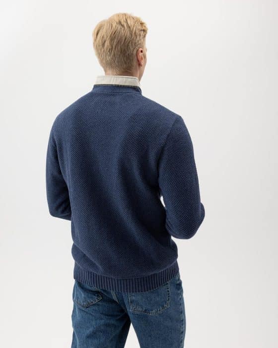 A person with short, blonde hair is standing and facing away. They are wearing a textured navy sweater over a light-colored collared shirt, paired with blue jeans. The background is plain white.