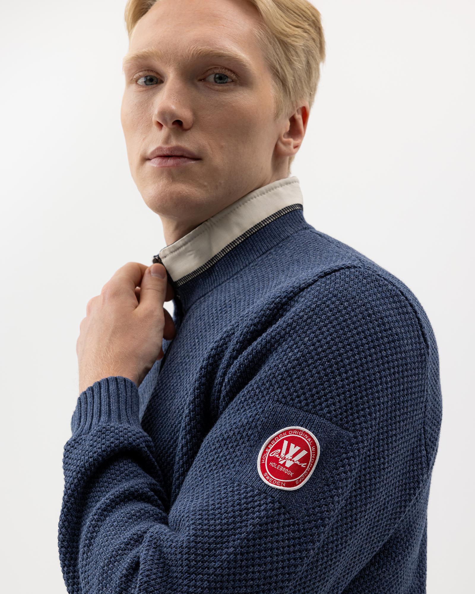 A person with blonde hair wearing a textured navy sweater and adjusting their grey collared shirt. The sweater has a red circular patch with white text on the sleeve. The background is plain white.