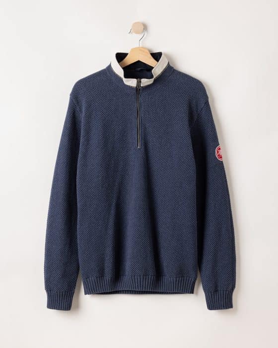 A navy blue textured pullover with a quarter-zip and light gray collar is hanging on a wooden hanger against a plain wall. The sleeve has a red and white circular patch.