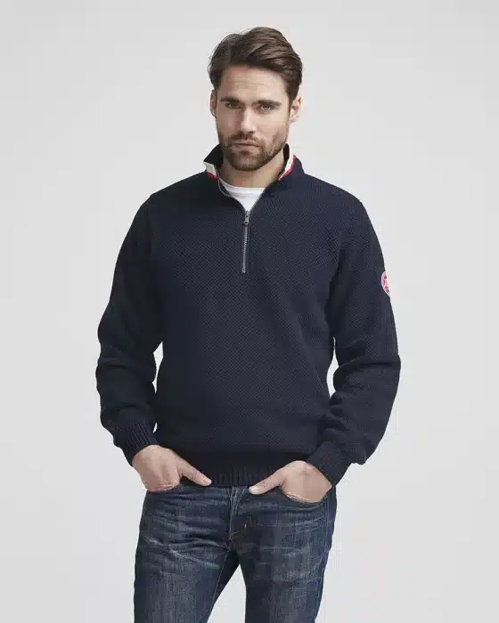 Man wearing a navy quarter-zip sweater with a subtle pattern and a patch on the left sleeve, paired with blue jeans. He stands against a plain white background, with his hands in his pockets, looking at the camera.