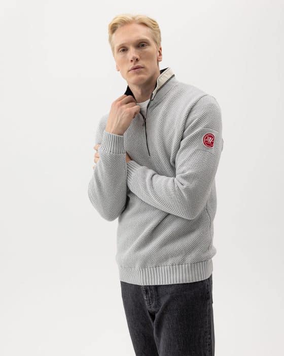 A person with short blond hair wears a light gray textured zip-up sweater with a red circular patch on the sleeve. The sweater is partially unzipped, revealing a beige interior. They are also wearing dark jeans against a plain white background.