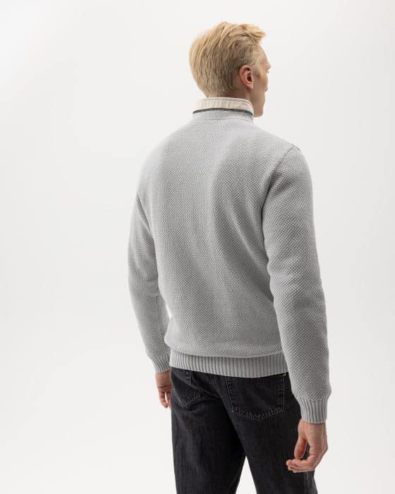 A person with light hair is shown from the back, wearing a light gray textured sweater over a white turtleneck, paired with dark jeans. The background is plain white.