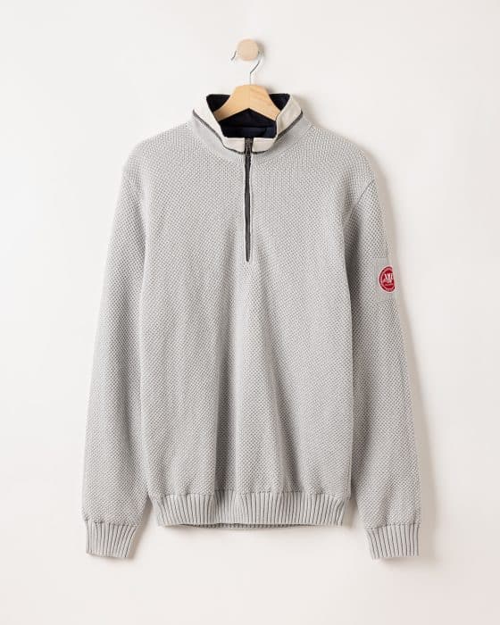 A light gray sweater with a textured pattern hangs on a wooden hanger against a cream wall. It features a quarter zip and a red circular patch on the left sleeve. The collar is slightly raised.