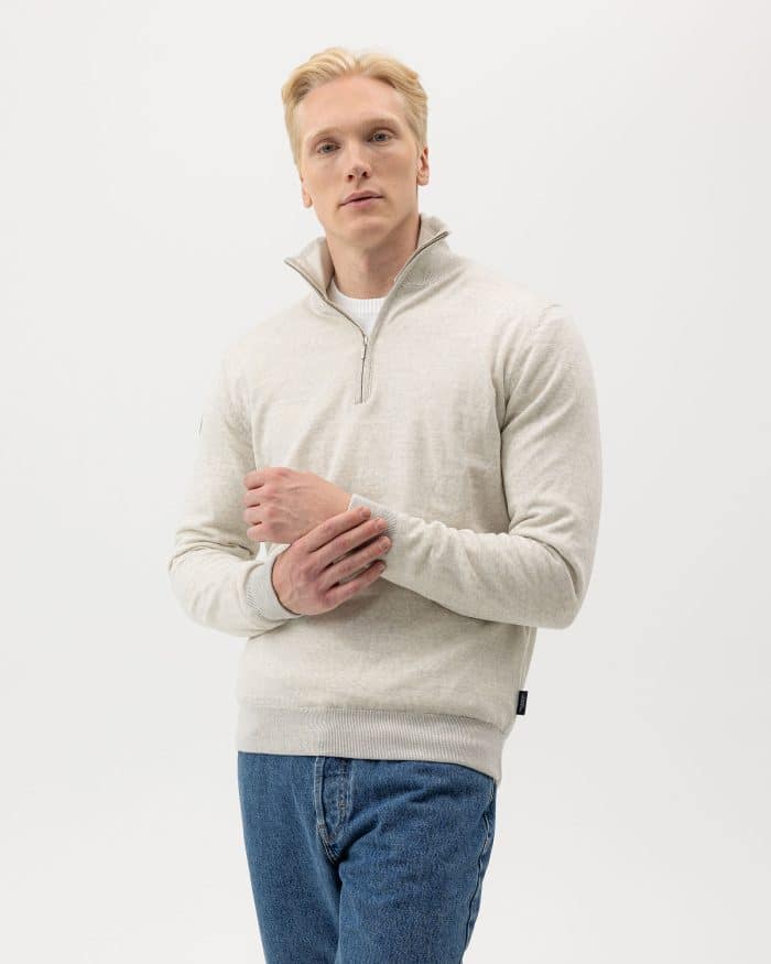 A person with short blonde hair is wearing a light gray quarter-zip pullover over a white shirt and blue jeans. They are standing against a plain white background, looking forward with one hand touching their other arm.