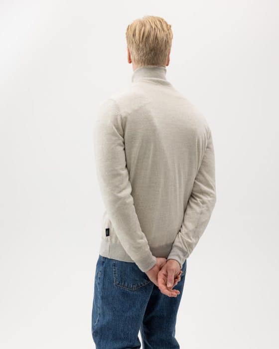 A person with blonde hair, wearing a light gray turtleneck sweater and blue jeans, stands with their back to the camera. Their hands are clasped behind their back. The background is plain white.