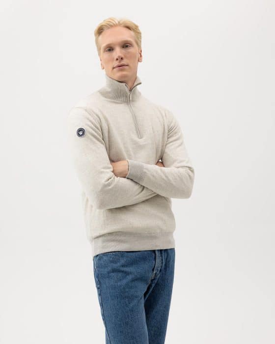 A person with light hair is wearing a light gray quarter-zip sweater and blue jeans. They are standing with arms folded against a plain white background. Theres a small circular patch on the upper sleeve of the sweater.