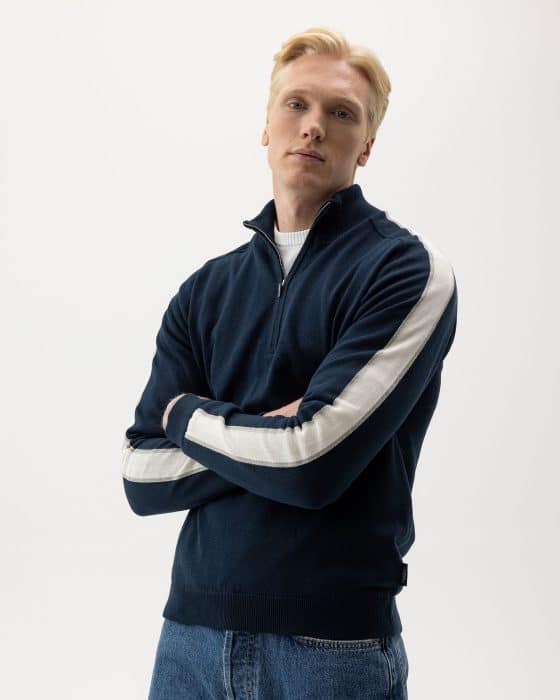 A person with short blonde hair wears a dark blue zip-up sweater with white stripes on the sleeves, paired with blue jeans. They stand against a plain white background with arms crossed, looking at the camera.