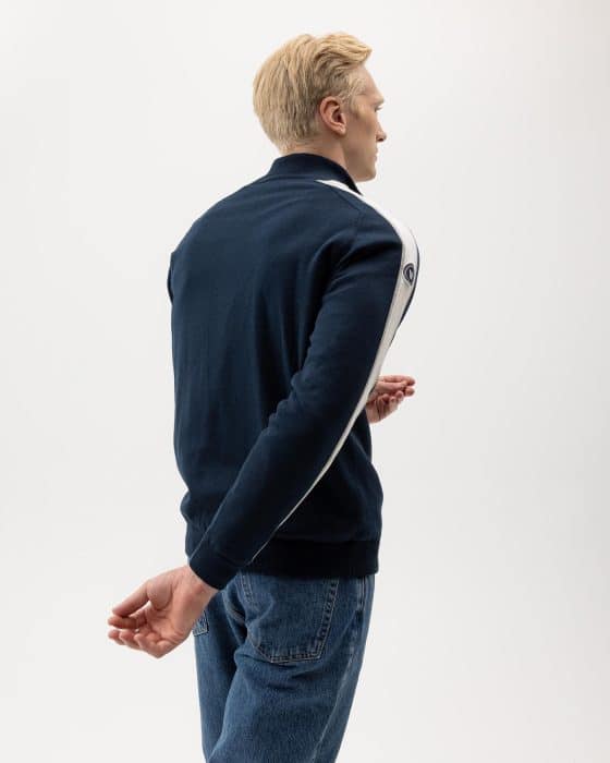 A person with short blond hair is standing against a light background. They are wearing a dark jacket with a white stripe on the sleeve and blue jeans. Their hands are slightly behind their back and one is clasping the other wrist.