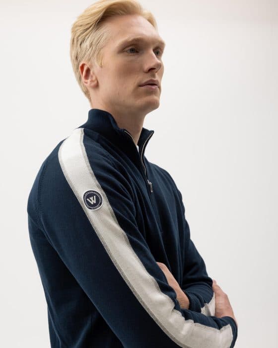 A person with short blond hair is wearing a dark blue jacket with white stripes on the sleeves. There is a circular logo on the upper arm. The individual is crossing their arms and gazing sideways against a plain white background.