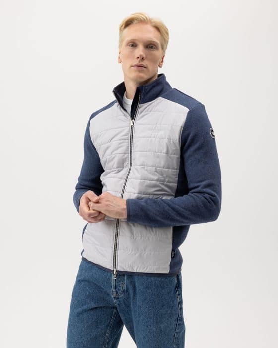 A person wearing a blue and gray zip-up jacket stands against a plain white background. The jacket has quilted front panels and dark sleeves. They are also wearing blue jeans and have short blond hair.