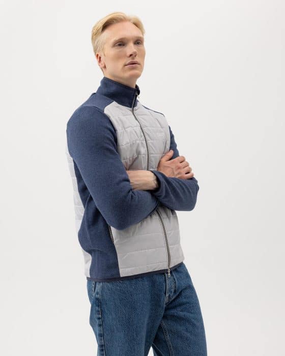 A person with light hair, wearing a blue and gray zip-up jacket and blue jeans, stands with arms crossed against a plain white background.