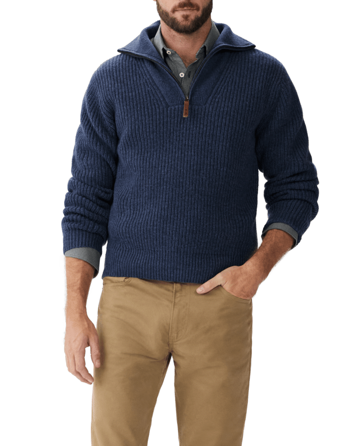 A man wearing a dark blue quarter-zip sweater over a collared shirt and tan pants stands with one hand in his pocket against a plain background.