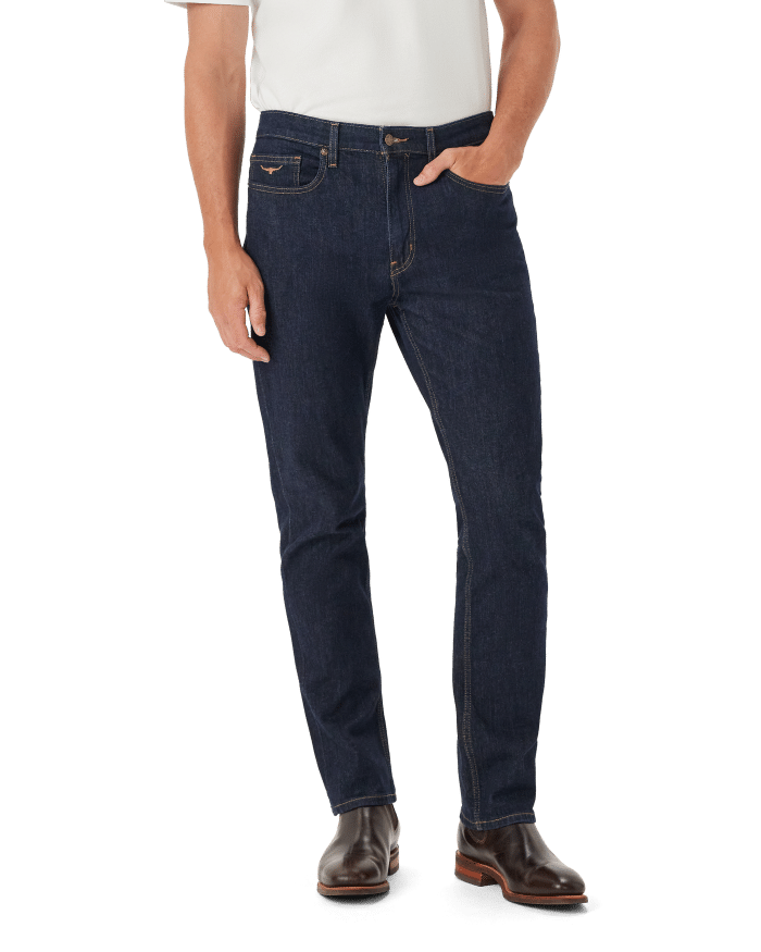 A person wearing a white shirt, dark blue slim-fit jeans, and brown boots stands with one hand in their pocket against a white background.