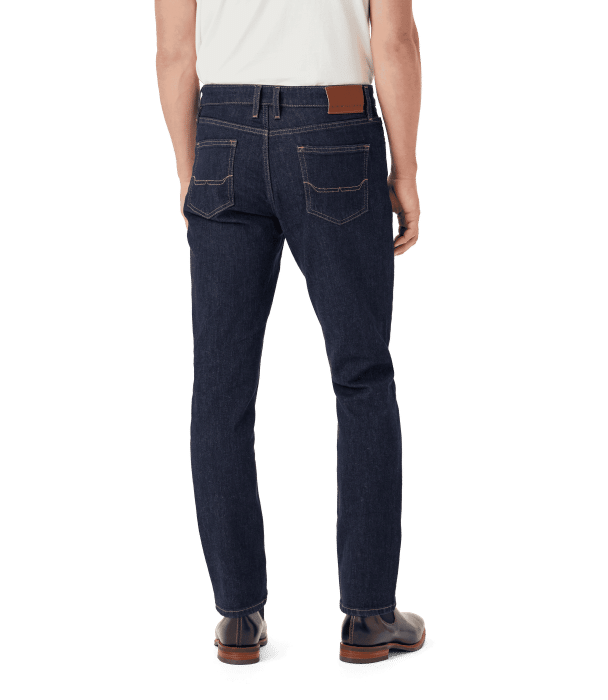 A person wearing a white shirt and dark blue jeans is standing facing away from the camera. The jeans have two back pockets, a brown patch on the waistband, and the person is wearing brown shoes.