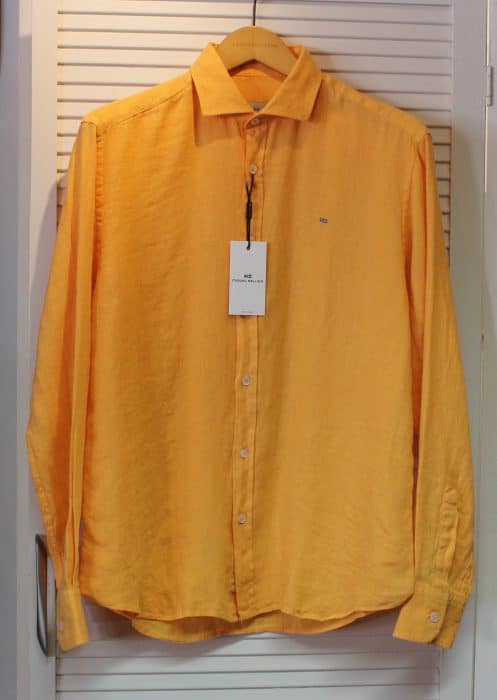A long-sleeved, button-up yellow shirt hangs on a wooden hanger. The shirt has a collar, a white tag attached to the top button, and is displayed against a white louvered background.