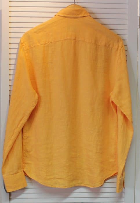 A long-sleeved, button-up yellow shirt is hanging on a hanger against a white louvered closet door. The shirt is displayed from the back.