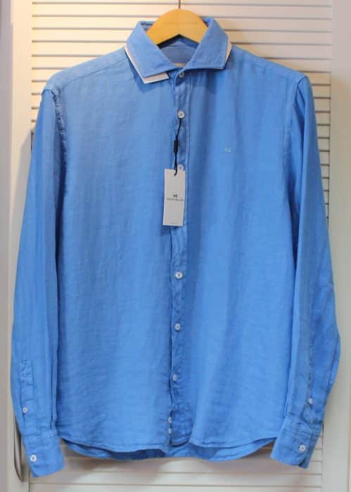 A light blue, long-sleeved button-up shirt with a white collar, hanging on a wooden hanger. A tag is attached to the top buttonhole, and the shirt is displayed against a white slatted background.