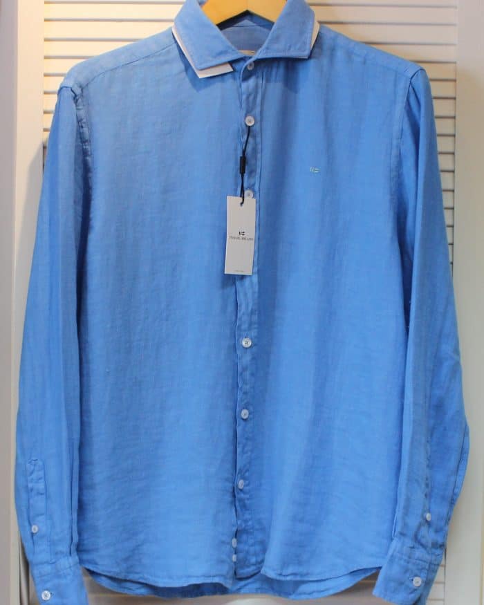 A light blue, long-sleeved button-up shirt with a white collar, hanging on a wooden hanger. A tag is attached to the top buttonhole, and the shirt is displayed against a white slatted background.