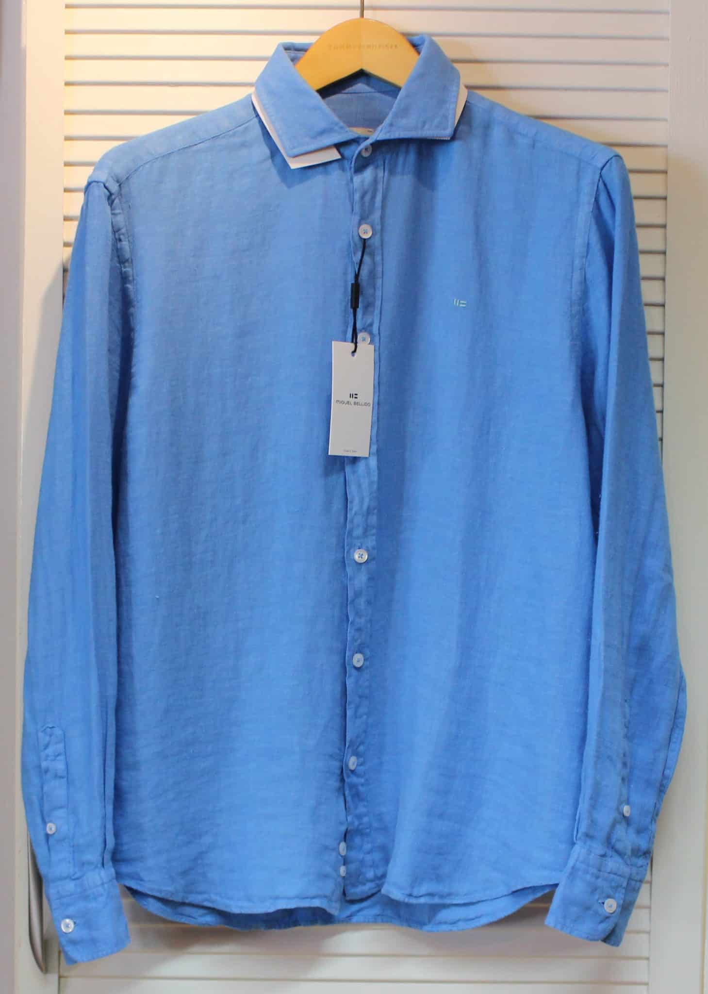 A light blue, long-sleeved button-up shirt with a white collar, hanging on a wooden hanger. A tag is attached to the top buttonhole, and the shirt is displayed against a white slatted background.