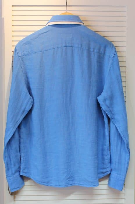 A long-sleeved, light blue button-up shirt is hanging on a wooden hanger against a white, slatted closet door. The shirt is displayed from the back.