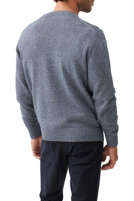 A man is shown from the back wearing a textured, light gray-blue sweater and dark pants. His head is turned slightly to the side, and his right arm is bent at the elbow. The background is white.