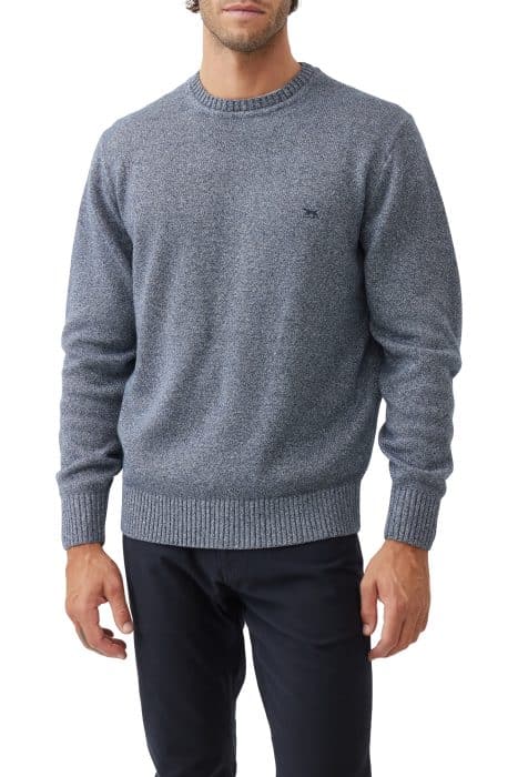 A man wearing a textured blue crewneck sweater with ribbed cuffs and hem, paired with dark pants, standing against a white background. His face is partially out of frame.