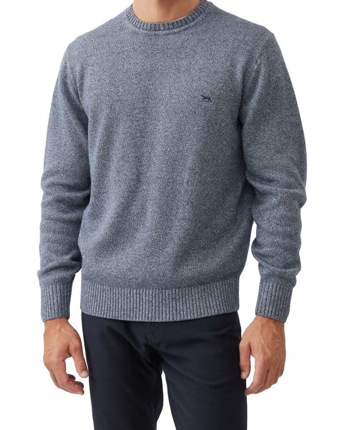 A man wearing a textured blue crewneck sweater with ribbed cuffs and hem, paired with dark pants, standing against a white background. His face is partially out of frame.