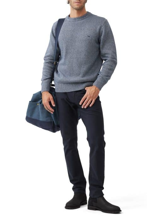 A man wearing a light grey sweater, dark blue pants, and black boots stands holding a blue duffel bag. He has a dark blue shoulder bag slung across his body. The background is white.