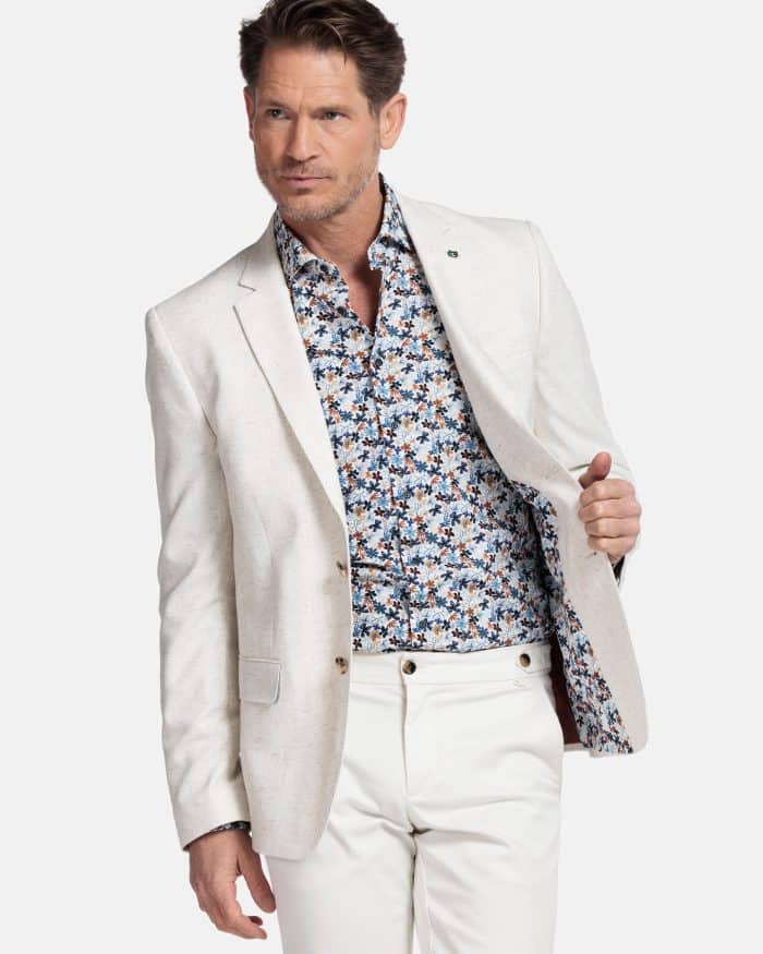 A man wearing a light beige blazer over a floral-patterned shirt and white pants stands against a plain white background, holding one side of his blazer open to reveal part of the shirt and blazer lining.