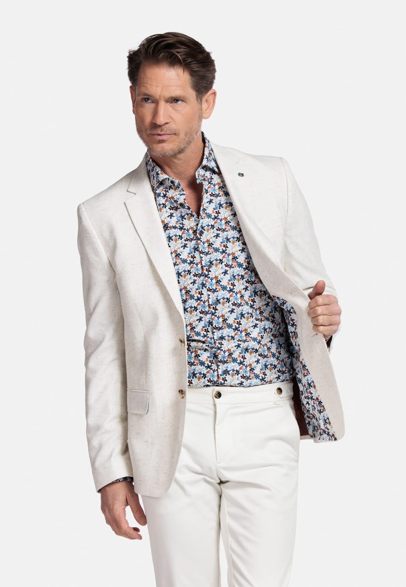 A man wearing a light beige blazer over a floral-patterned shirt and white pants stands against a plain white background, holding one side of his blazer open to reveal part of the shirt and blazer lining.