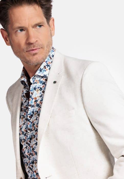A man with styled brown hair and light stubble wears a white blazer over a floral-patterned shirt, looking to his left against a plain light background.