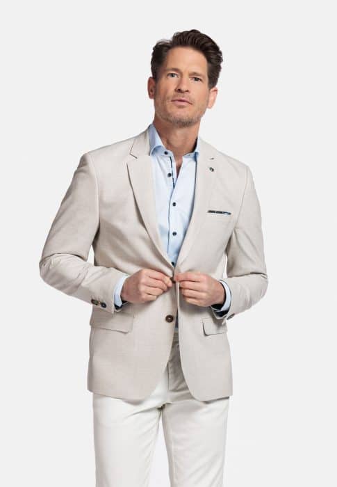 A man wearing a light beige blazer over a light blue dress shirt and white pants stands against a plain white background, looking slightly to the side with a neutral expression.