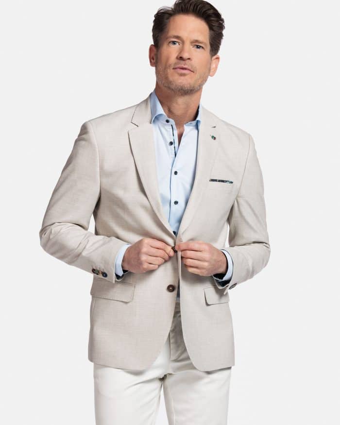 A man wearing a light beige blazer over a light blue dress shirt and white pants stands against a plain white background, looking slightly to the side with a neutral expression.