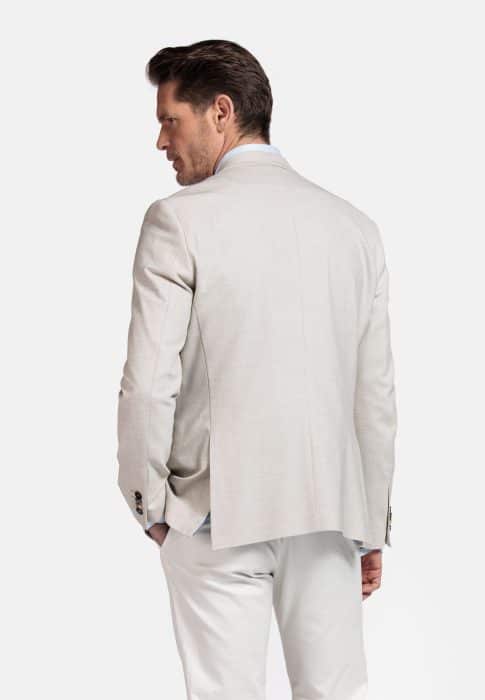A man with short brown hair wears a light beige blazer and white pants, standing with his back partially turned to the camera and his hands in his pockets against a plain white background.