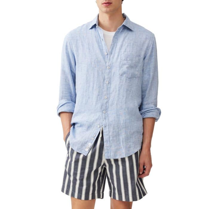 A person wearing a light blue button-up linen shirt over a white T-shirt and black-and-white vertically striped shorts, standing with one hand in their pocket against a plain white background.