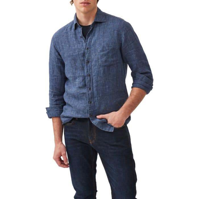 A man wearing a long-sleeve, blue button-up shirt over a black T-shirt and dark jeans, standing with one hand in his pocket against a white background.