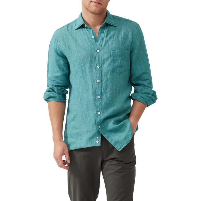 A man wearing a teal long-sleeve button-up shirt with rolled-up sleeves and dark pants, standing with one hand in his pocket against a plain white background.