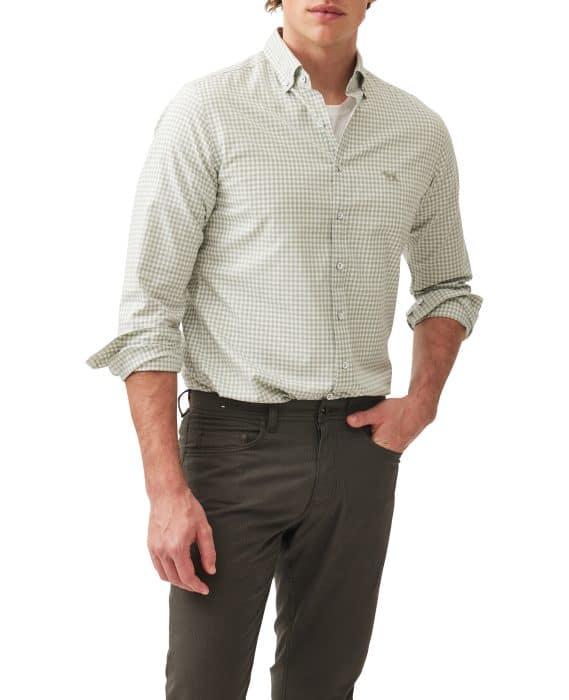 A man wearing a light green and white checkered button-down shirt with rolled-up sleeves, dark gray pants, and his left hand in his pocket, standing against a plain white background.