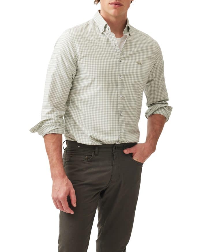A man wearing a light green and white checkered button-down shirt with rolled-up sleeves, dark gray pants, and his left hand in his pocket, standing against a plain white background.