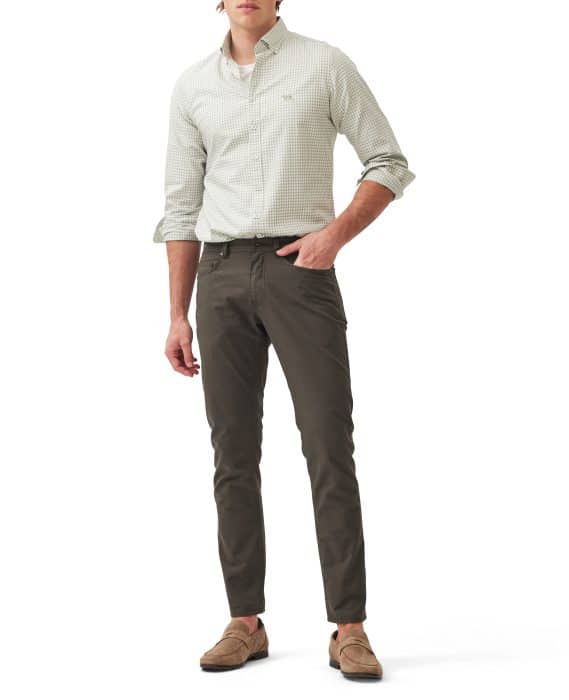 A man stands wearing a light green checkered button-up shirt with rolled-up sleeves, dark slim-fit pants, and light brown loafers. His left hand is in his pocket and his right arm rests at his side.