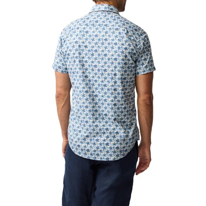 A man is seen from behind wearing a short-sleeve, button-up shirt with a blue and white geometric leaf pattern and dark blue pants, standing against a white background.