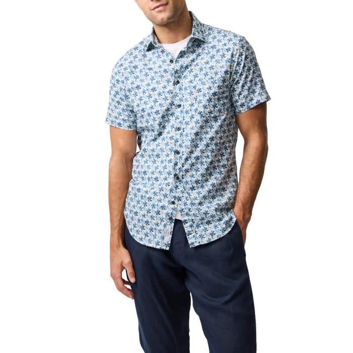 A man wearing a short-sleeve, button-up shirt with a blue and white floral pattern, dark blue pants, and a white undershirt, standing with one hand in his pocket against a white background.