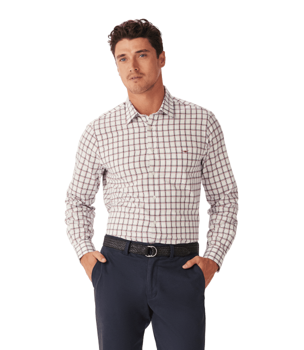 A man wearing a long-sleeve, white button-up shirt with a red and blue checkered pattern and dark blue pants stands with his hands in his pockets, looking forward.