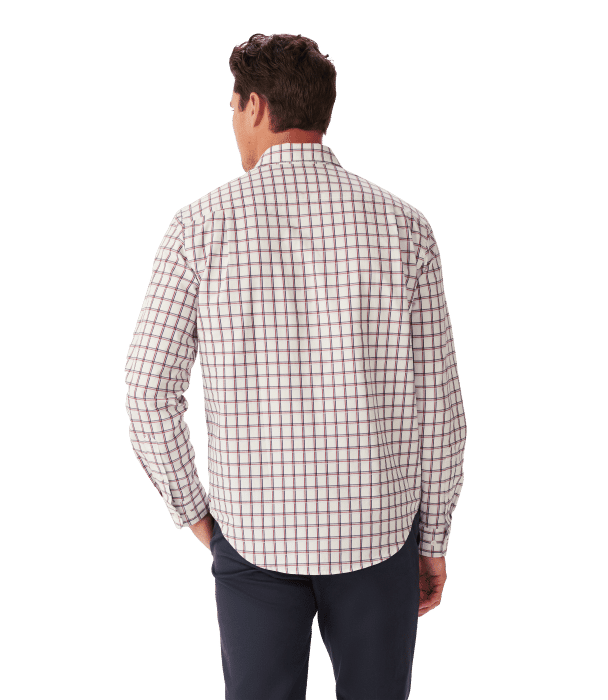 A man stands facing away, wearing a long-sleeve, white button-up shirt with a red and blue plaid pattern and dark pants. His left hand is in his pocket.